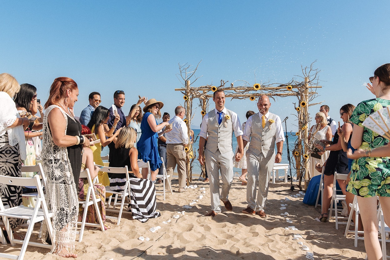 Two grooms walking down the aisle at a beach wedding ceremony with sunflower arch